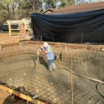 A construction site showing a backyard pool being built, with workers and equipment visible around the site.