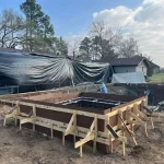 A construction site showing a backyard pool being built, with workers and equipment visible around the site.