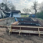 A construction site showing a backyard pool being built, with workers and equipment visible around the site.