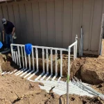Two men constructing a wooden fence in a dirt area, focused on their task with tools in hand.