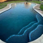 A backyard pool filled with clear blue water, surrounded by green grass and a few decorative plants.